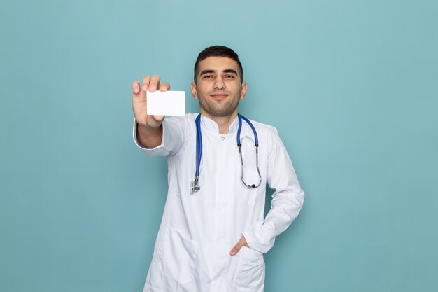 Vista frontal de um jovem médico de terno branco com estetoscópio azul segurando um cartão branco