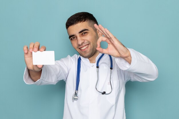 Vista frontal de um jovem médico de terno branco com estetoscópio azul segurando um cartão branco