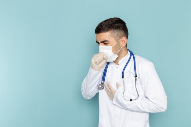 Vista frontal de um jovem médico de terno branco com estetoscópio azul usando máscara estéril tossindo