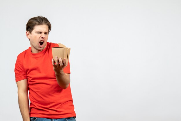 Vista frontal de um jovem nervoso e zangado com uma blusa vermelha, segurando uma pequena caixa no fundo branco