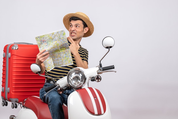 Vista frontal de um jovem perplexo com um chapéu de palha na motocicleta segurando o mapa