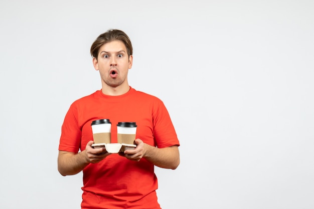 Vista frontal de um jovem preocupado com uma blusa vermelha segurando café em copos de papel no fundo branco