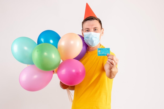 Vista frontal de um jovem segurando balões coloridos e um cartão do banco na máscara na parede branca