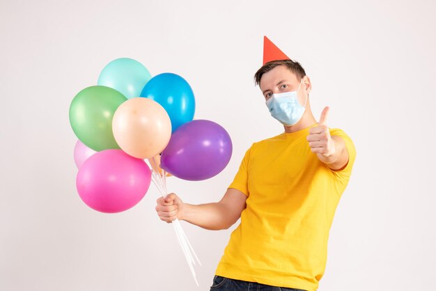 Vista frontal de um jovem segurando balões coloridos em uma máscara estéril na parede branca