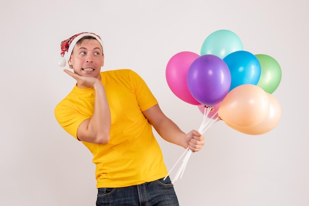 Vista frontal de um jovem segurando balões coloridos na parede branca