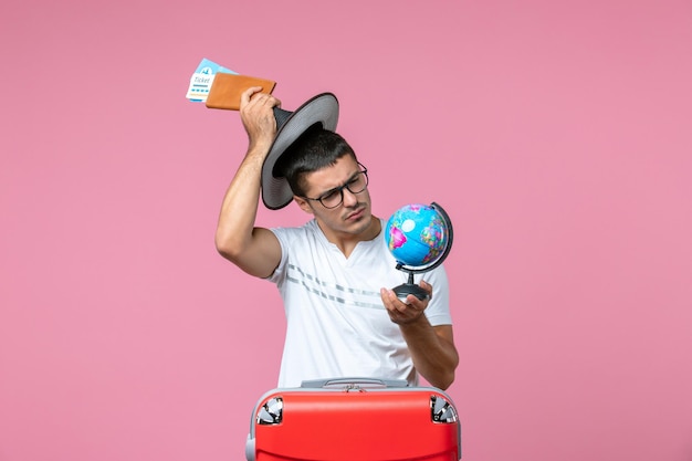 Vista frontal de um jovem segurando os bilhetes de férias e o globo na parede rosa