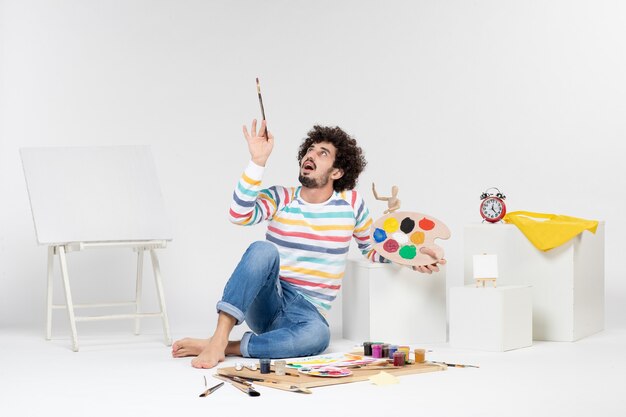 Vista frontal de um jovem segurando tintas e borla para desenhar na parede branca