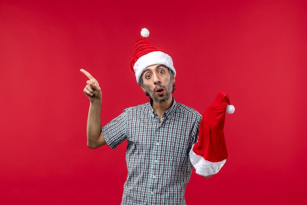 Foto grátis vista frontal de um jovem segurando um boné vermelho de natal na parede vermelha