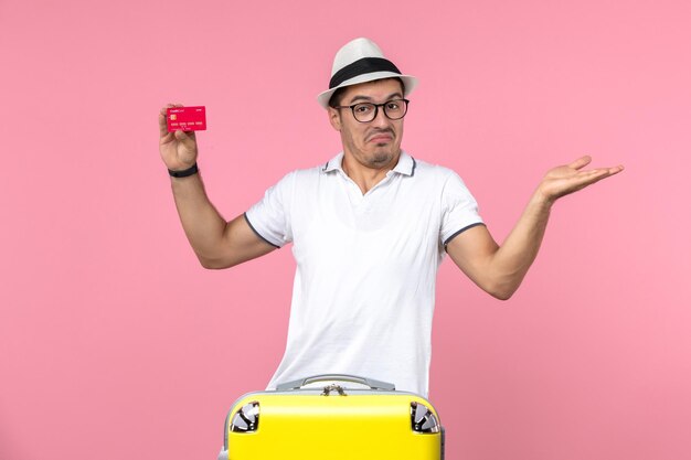 Vista frontal de um jovem segurando um cartão de banco vermelho de férias na parede rosa