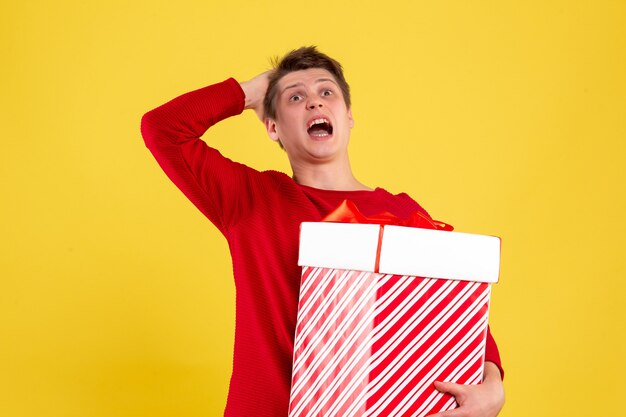 Vista frontal de um jovem segurando um grande presente de Natal na parede amarela