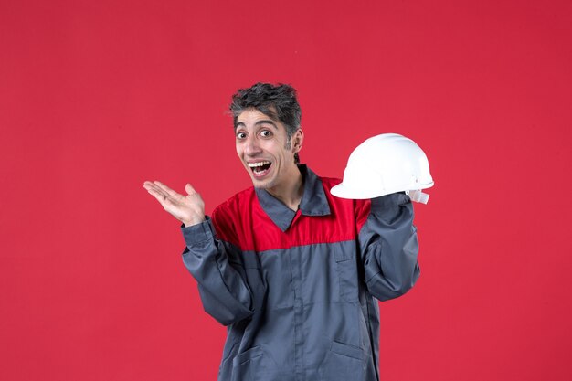 Vista frontal de um jovem trabalhador feliz de uniforme e segurando um capacete apontando algo do lado direito na parede vermelha isolada