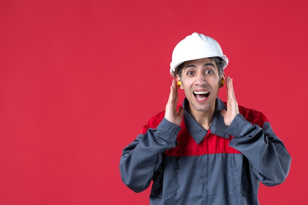 Vista frontal de um jovem trabalhador satisfeito e sorridente, de uniforme, com capacete e tampões de ouvido na parede vermelha isolada