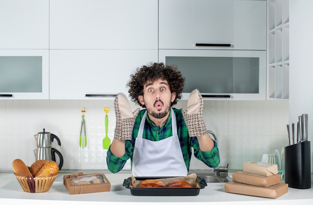 Vista frontal de um jovem usando o suporte em pé atrás de uma mesa de massa recém-assada na cozinha branca