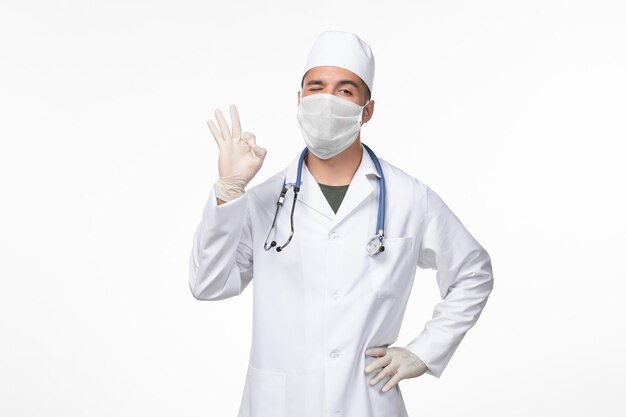 Vista frontal de um médico com uniforme médico e máscara contra cobiça com estetoscópio piscando no vírus da parede branca - doença pandêmica