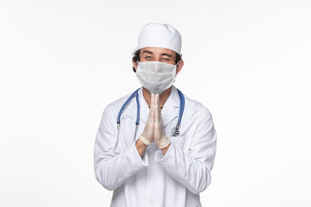 Vista frontal de um médico em terno médico usando máscara estéril como proteção contra a oração cobiçosa sobre doença pandêmica de coronavírus de vírus de parede branca