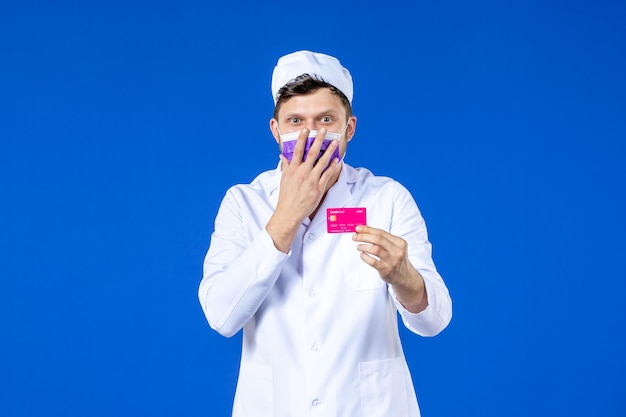 Vista frontal de um médico em traje médico e máscara segurando um cartão de crédito em azul