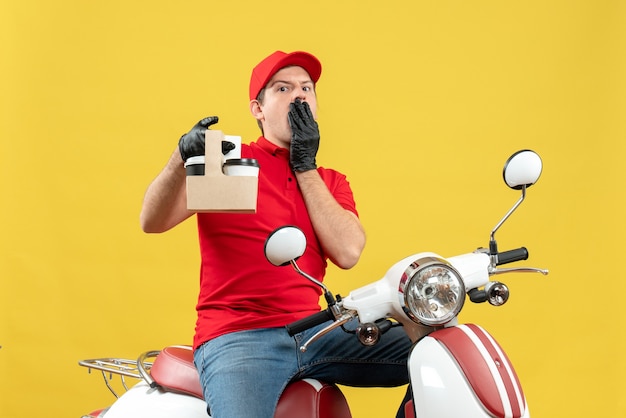 Vista frontal de um mensageiro emocionalmente chocado, vestindo uma blusa vermelha e luvas de chapéu, com máscara médica, entregando pedidos sentado na scooter segurando pedidos