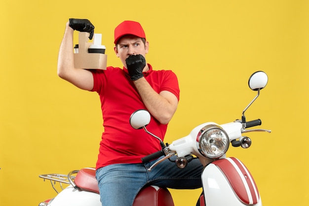 Vista frontal de um mensageiro surpreso usando blusa vermelha e luvas de chapéu com máscara médica, entregando pedidos sentado na scooter segurando pedidos