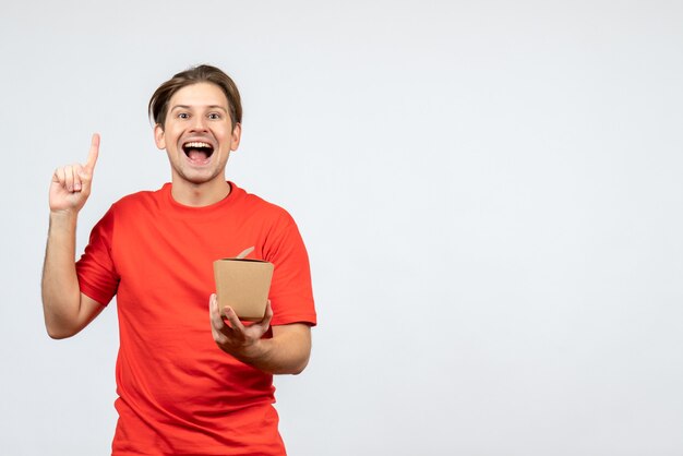 Vista frontal de um rapaz sorridente com uma blusa vermelha, segurando uma pequena caixa e apontando para cima no fundo branco