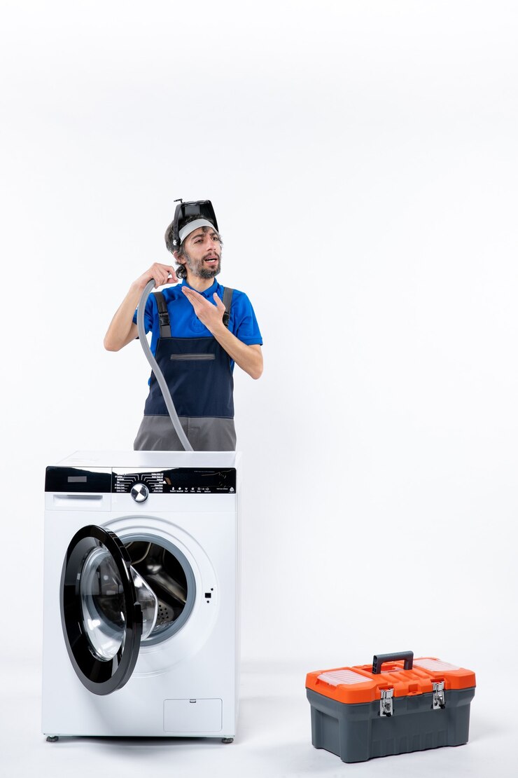 Foto grátis vista frontal de um reparador confuso de uniforme em pé atrás da máquina de lavar, segurando um tubo de plástico na parede branca