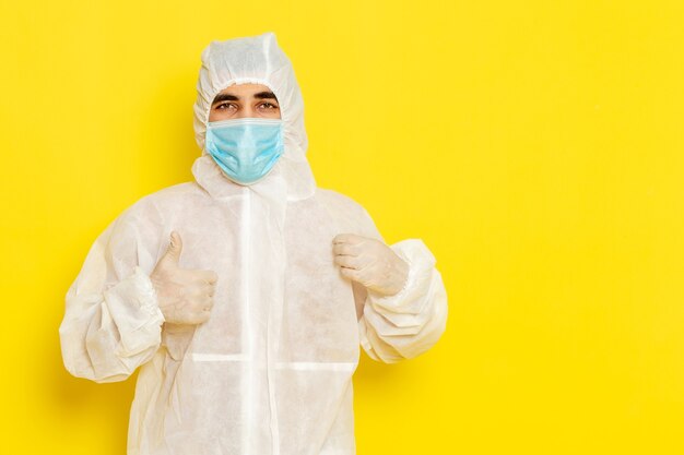 Vista frontal de um trabalhador científico do sexo masculino em traje de proteção especial e com máscara posando na parede amarela clara
