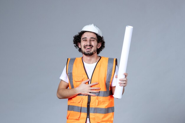 Vista frontal de um trabalhador da construção civil sorridente em colete de advertência com capacete de segurança e segurando em branco na parede cinza