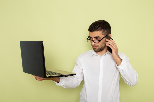 Vista frontal de um trabalhador de escritório com camisa branca segurando um laptop falando ao telefone na parede verde