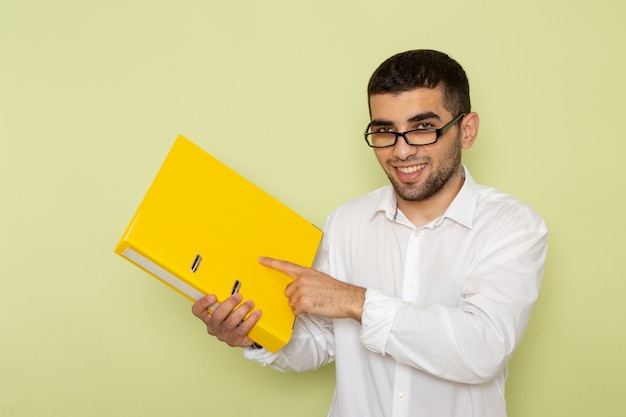Vista frontal de um trabalhador de escritório com camisa branca segurando uma pasta amarela na parede verde-clara