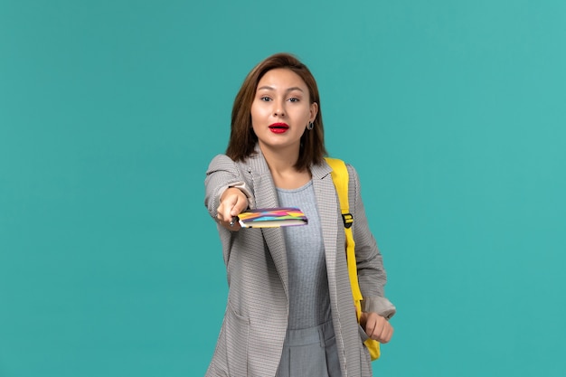 Vista frontal de uma aluna com uma jaqueta cinza e uma mochila amarela segurando um caderno na parede azul