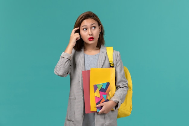 Vista frontal de uma aluna com uma jaqueta cinza usando uma mochila amarela segurando arquivos e o caderno afinando na parede azul