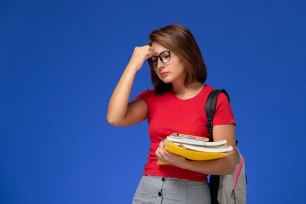 Vista frontal de uma aluna de camisa vermelha com uma mochila segurando livros e arquivos deprimidos na parede azul