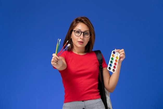 Vista frontal de uma aluna de camisa vermelha com uma mochila segurando tintas para desenho e borlas na parede azul