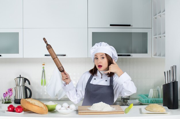 Foto grátis vista frontal de uma chef feminina confiante de uniforme em pé atrás da mesa com alimentos da tábua de cortar, segurando o rolo de massa, fazendo um gesto de me chamar na cozinha branca