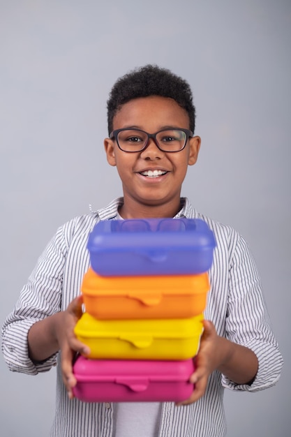 Vista frontal de uma criança sorridente em óculos mostrando caixas plásticas coloridas diante da câmera