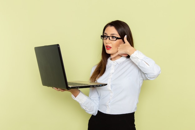 Vista frontal de uma funcionária de escritório com camisa branca e saia preta, usando seu laptop na parede verde-clara