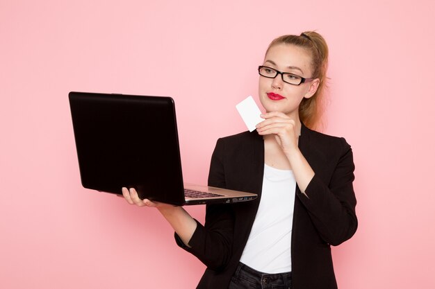 Vista frontal de uma funcionária de escritório com jaqueta preta, segurando um cartão branco e usando o laptop na parede rosa