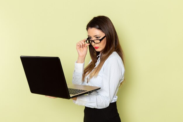 Vista frontal de uma funcionária de escritório de camisa branca e saia preta segurando o laptop na parede verde-clara