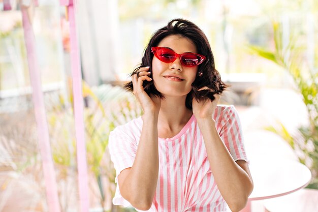 Vista frontal de uma garota europeia deslumbrante em camiseta listrada Mulher sorridente em óculos de sol rosa tocando cabelo no fundo desfocado