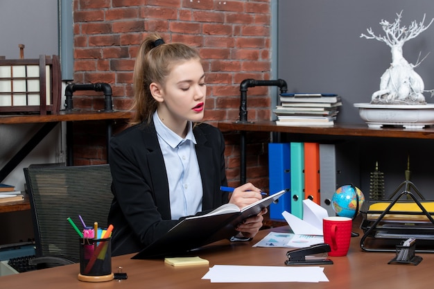 Foto grátis vista frontal de uma jovem assistente confiante sentada em sua mesa e escrevendo seu documento no escritório