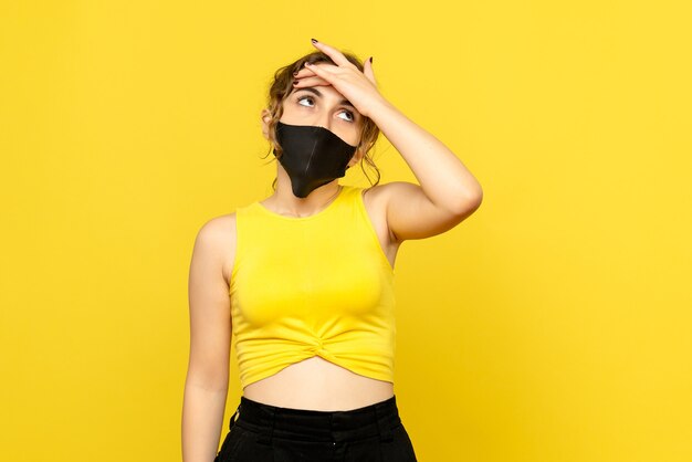 Vista frontal de uma jovem com máscara preta na parede amarela