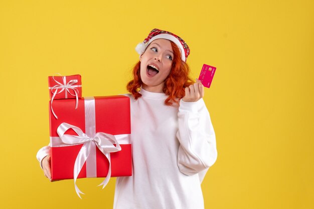 Vista frontal de uma jovem com presentes de Natal e cartão do banco na parede amarela