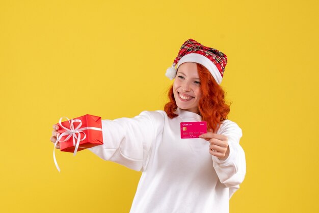 Vista frontal de uma jovem com um presente de Natal e cartão do banco na parede amarela
