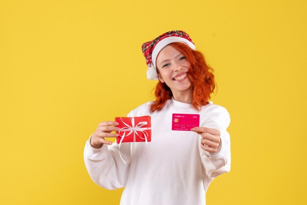 Vista frontal de uma jovem com um presente de Natal e cartão do banco na parede amarela