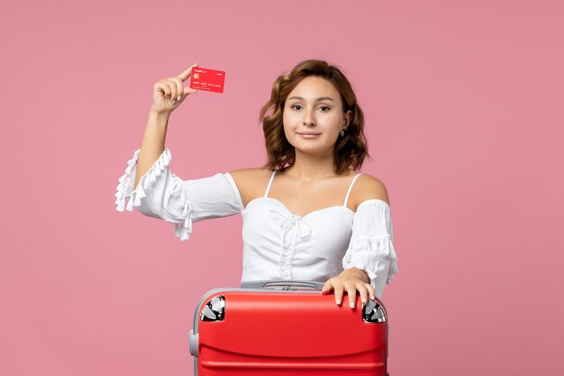 Vista frontal de uma jovem com uma sacola de férias segurando um cartão vermelho do banco na parede rosa