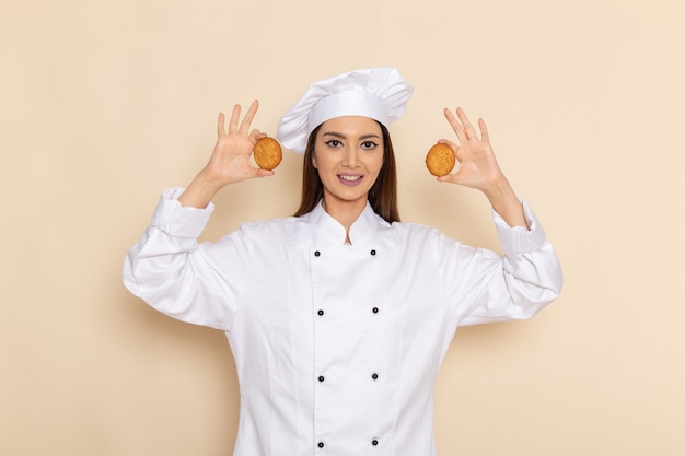 Vista frontal de uma jovem cozinheira em um terno branco segurando biscoitos com um sorriso na parede branca