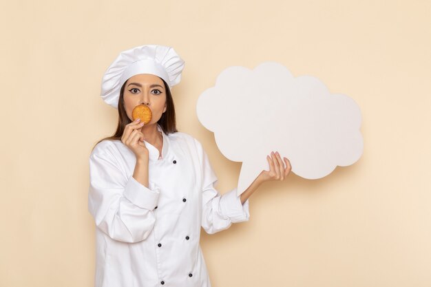 Foto grátis vista frontal de uma jovem cozinheira em um terno branco segurando um biscoito e uma placa branca na parede branca