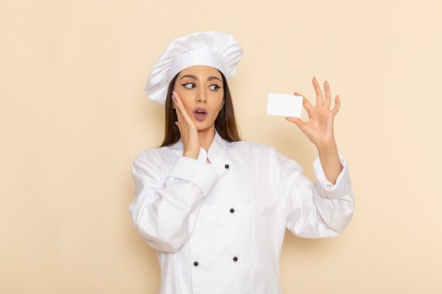 Vista frontal de uma jovem cozinheira em um terno branco, segurando um cartão branco na mesa branca-clara, cozinhando trabalho cozinha trabalho cozinha trabalhador