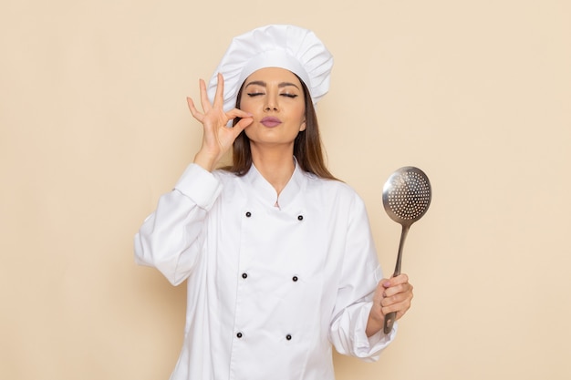 Vista frontal de uma jovem cozinheira em um terno branco segurando uma colher de prata grande na parede branca