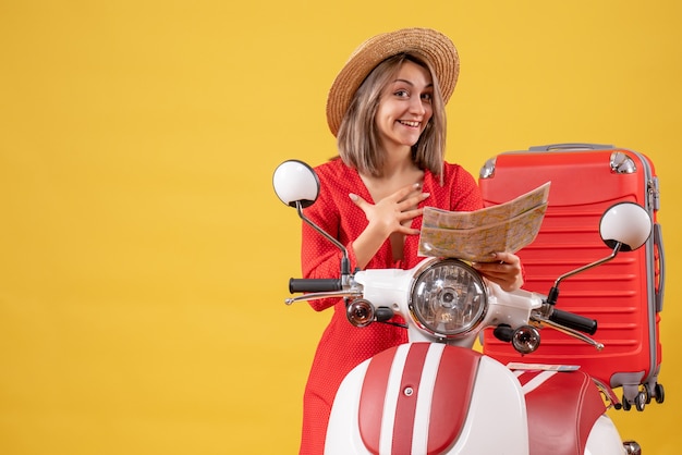 Vista frontal de uma jovem feliz com um vestido vermelho perto de um ciclomotor segurando um mapa