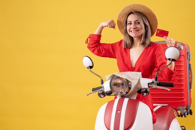 Vista frontal de uma jovem feliz em um vestido vermelho segurando um cartão de crédito, mostrando o músculo do braço perto de uma motocicleta